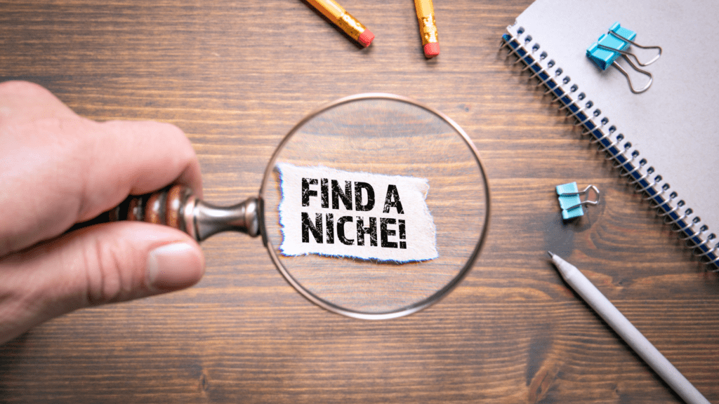 Magnifying glass focusing on the text 'Find a Niche!' next to a pencil and notepad with paper clips.
