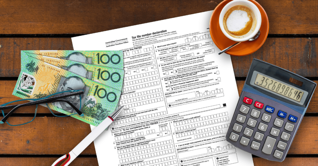 Top view of Australian tax forms, $100 notes, a calculator, coffee, and glasses on a wooden table.
