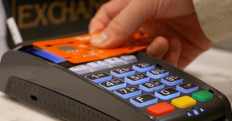 A close-up of a person's hand inserting an orange credit card into a payment terminal with illuminated keypad buttons.