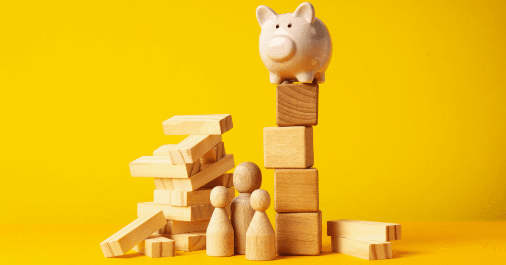 Wooden figures and blocks arranged in a strategic manner with a piggy bank perched atop, all set against a vibrant yellow background.