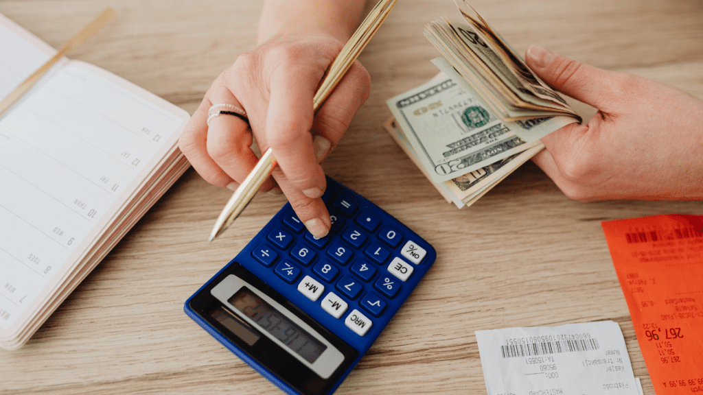 Person calculating finances with a calculator, cash, and planner on a desk.